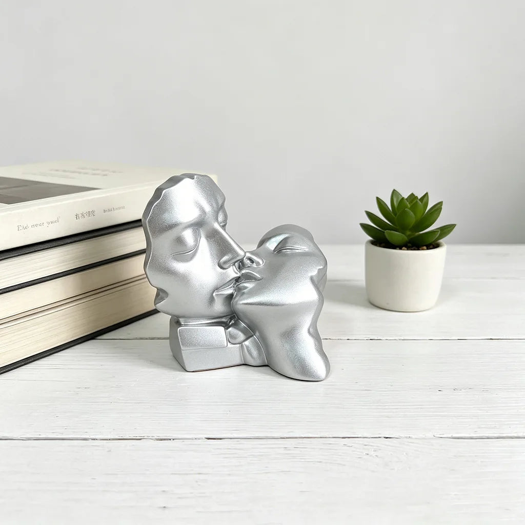 Silver resin sculpture of a kissing couple resting on a white wooden surface next to a potted succulent plant.