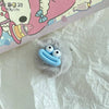 A small, light blue sausage mouth charm made of felt or silicone with two white eyes, resting on crumpled white paper.