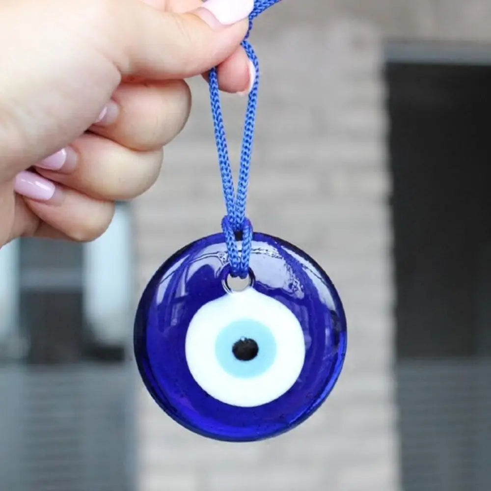 A hand holds a blue glass evil eye amulet pendant with a white iris and black pupil, suspended by a blue cord.