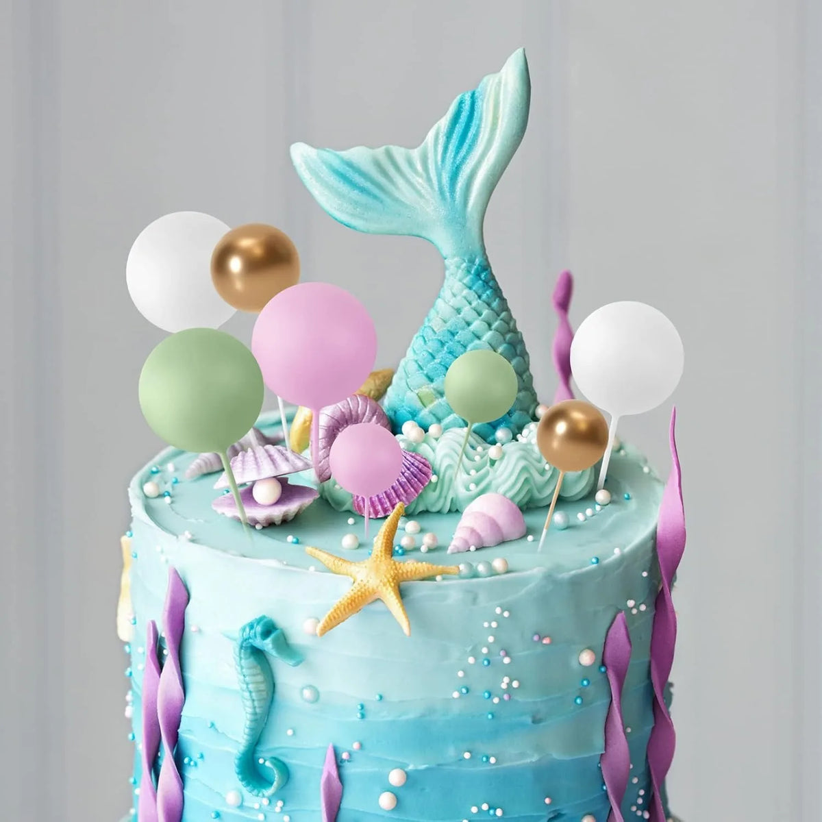 A blue cake decorated with sea creatures and a collection of light blue, pink, and gold foam ball toppers.