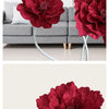 A close-up of deep red velvet artificial peony flowers displayed on white stems in a modern living room setting.