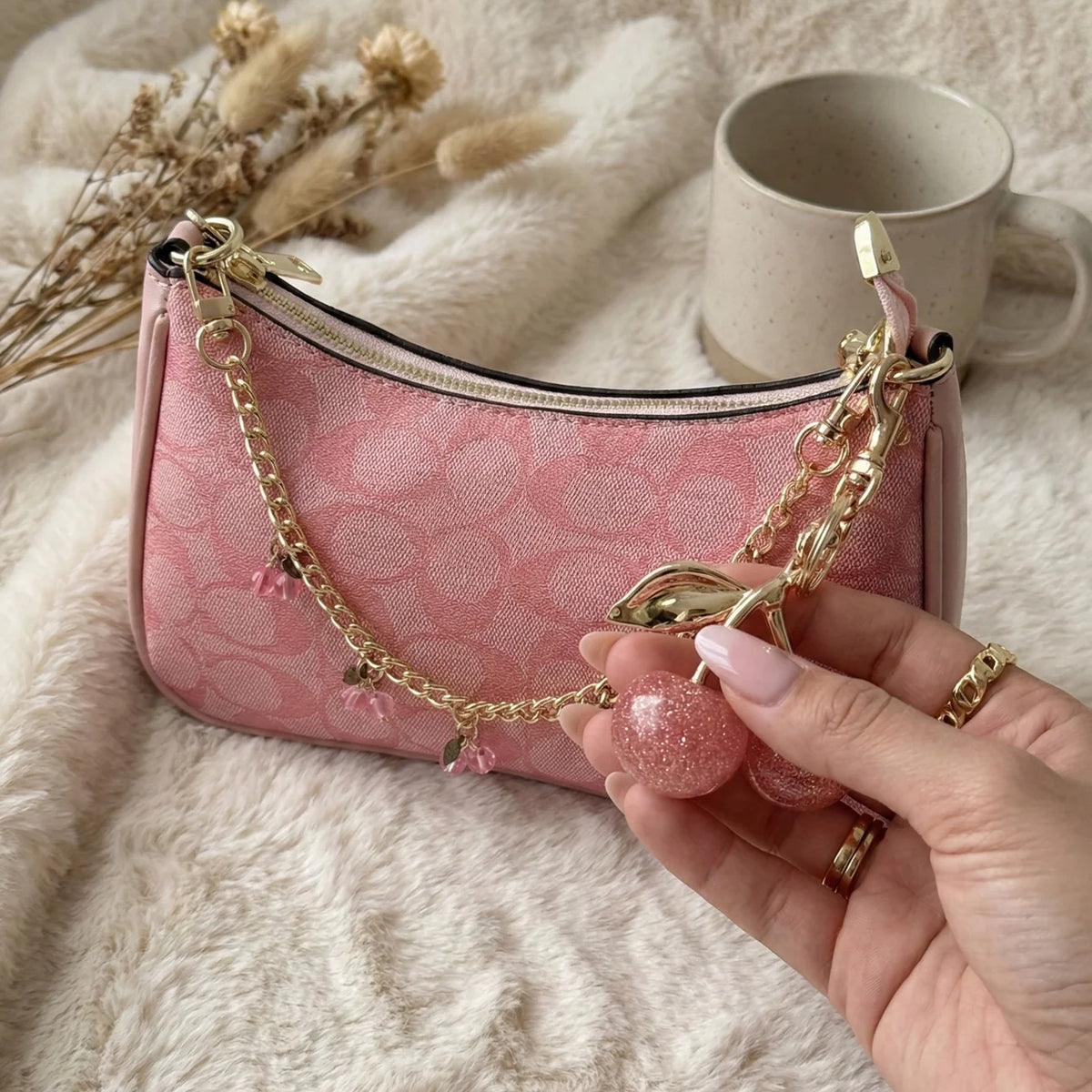 A pink velvet pouch with a gold chain and a pink glass stone charm is held in a hand on a fluffy white surface.