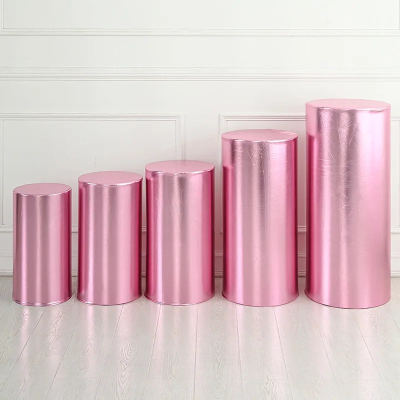 A row of four metallic pink spandex cylinder stands displayed against a white paneled wall.