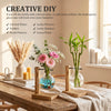 Installation DIY créative avec des vases en verre remplis d'eau colorée, de galets et de fleurs sur une table en bois.