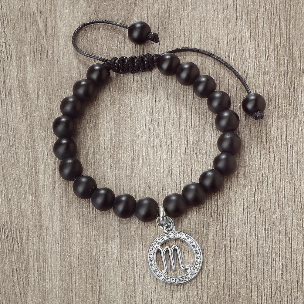 A black beaded bracelet with a silver zodiac sign charm is displayed on a wooden surface.