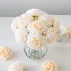 A bouquet of soft peach and cream silk peony artificial flowers in a glass vase on a white surface.