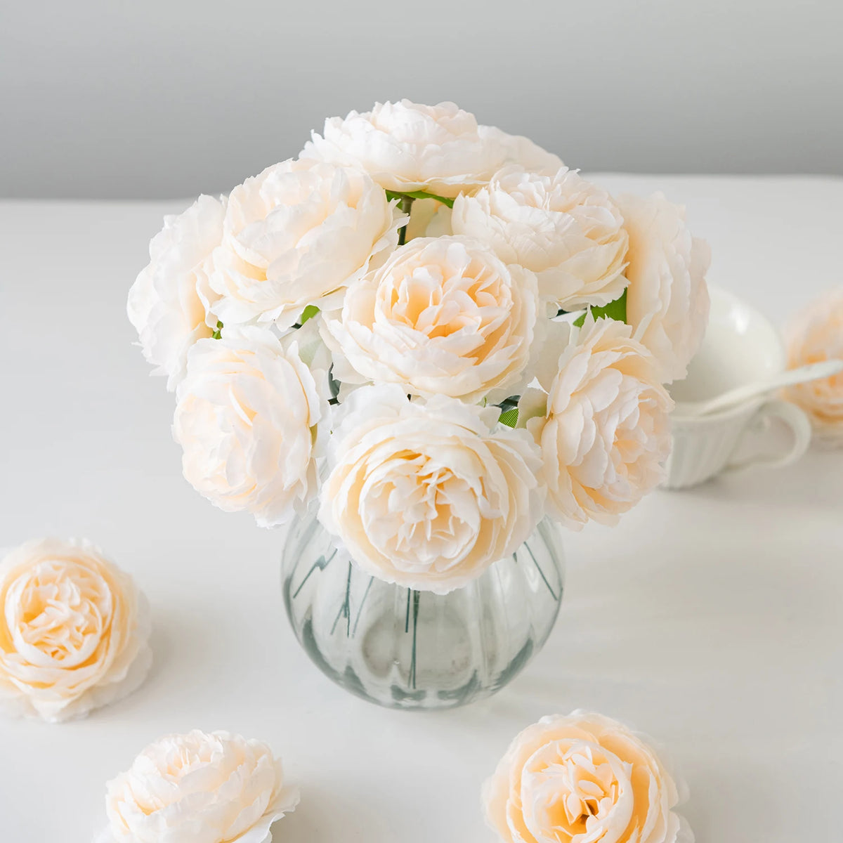 A bouquet of soft peach and cream silk peony artificial flowers in a glass vase on a white surface.
