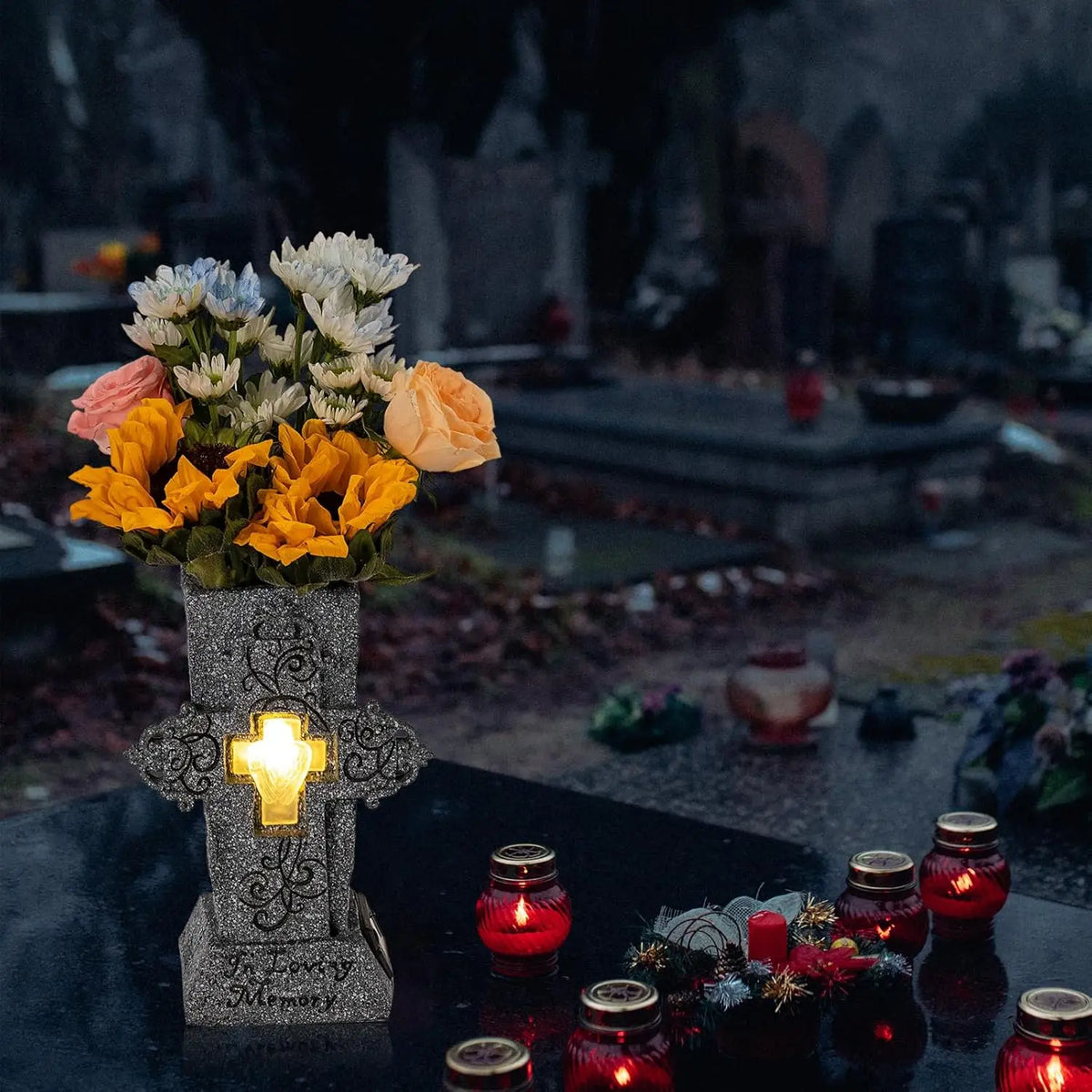 A dark stone vase with an illuminated cross holds a floral arrangement on a grave plot.