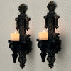A set of two vintage wrought iron wall-mounted candle holders with black bases and lit candles.