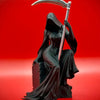 Schwarze Statue einer weiblichen Figur in fließender gotischer Kleidung, die eine große Sense vor einem roten Hintergrund häl