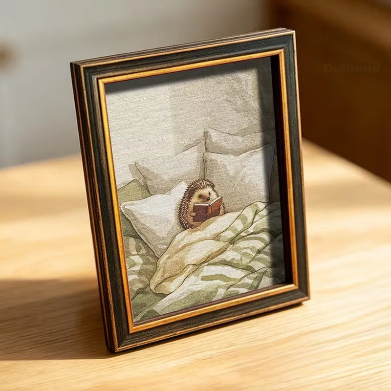 A framed photo of a small hedgehog sleeping under white bedding, displayed on a wooden surface.