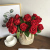 A bouquet of deep red artificial roses in a woven basket next to a white cup and framed artwork.