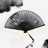 A black vintage bamboo folding hand fan with delicate floral designs is open against a bright sky.