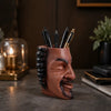 A terracotta-colored artistic face planter pot holds several black and gold pens on a dark surface.