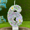 A number cake candle shaped like the number 6 covered in purple and green glitter with white flowers and a butterfly detail.