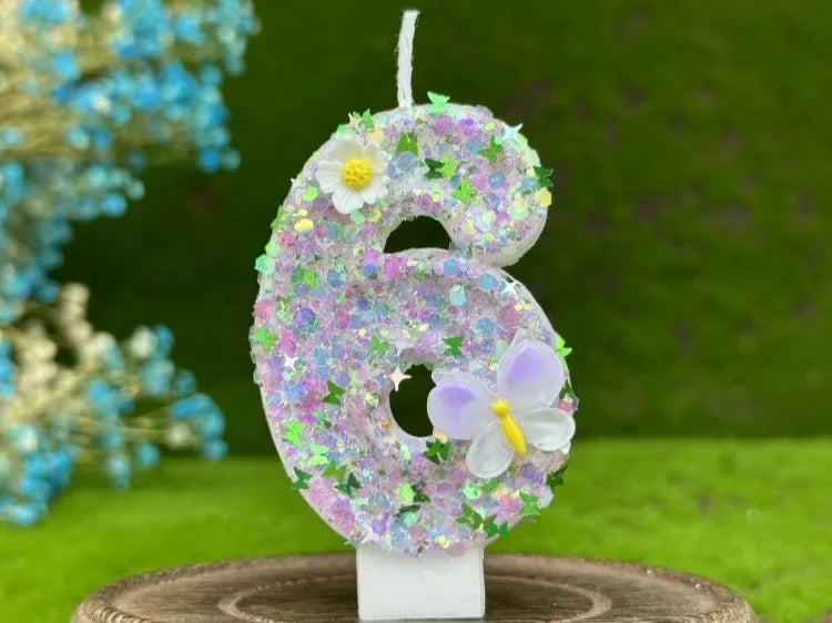 A number cake candle shaped like the number 6 covered in purple and green glitter with white flowers and a butterfly detail.