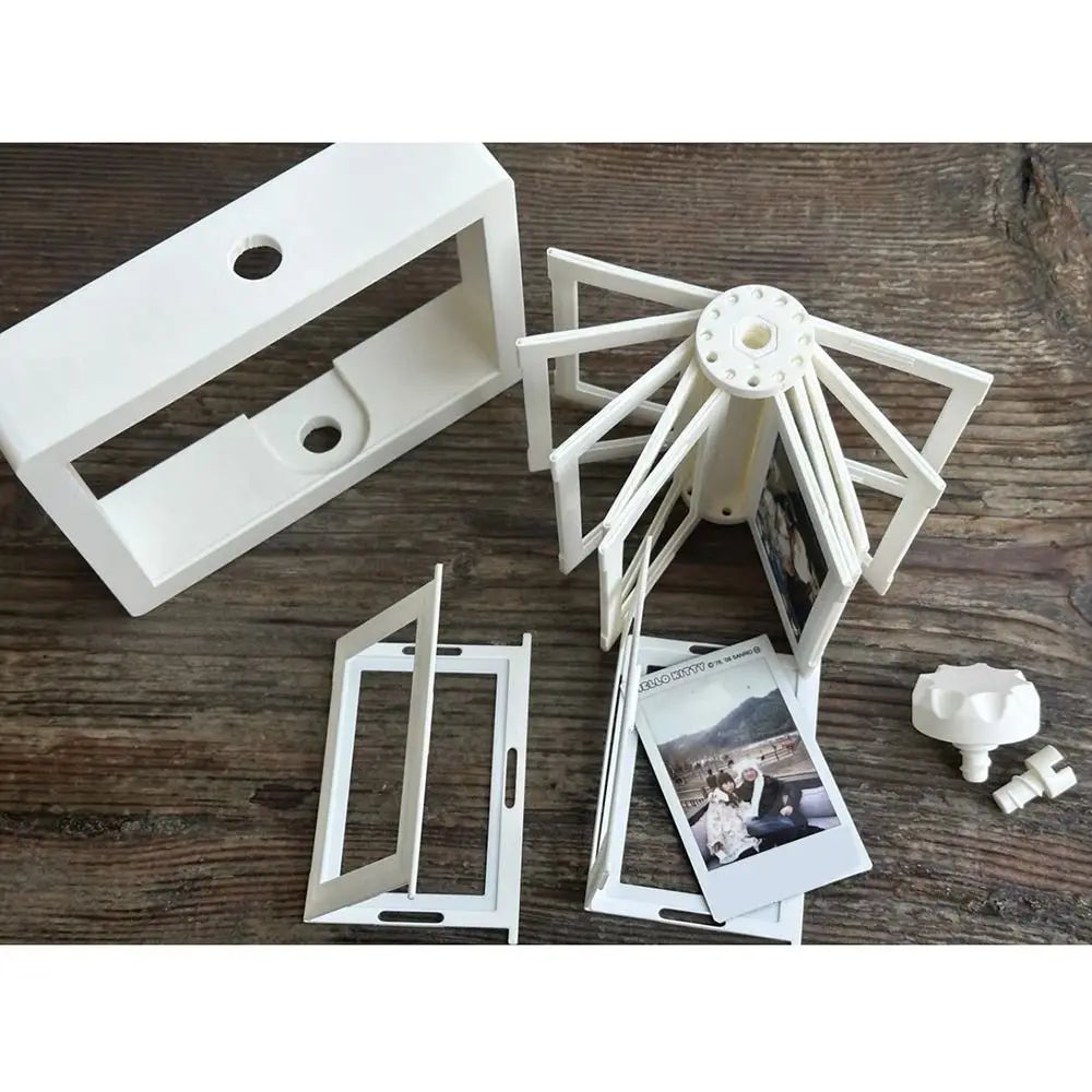 White 3D rotating photo holder with twelve clear display pockets shown on a wooden surface.
