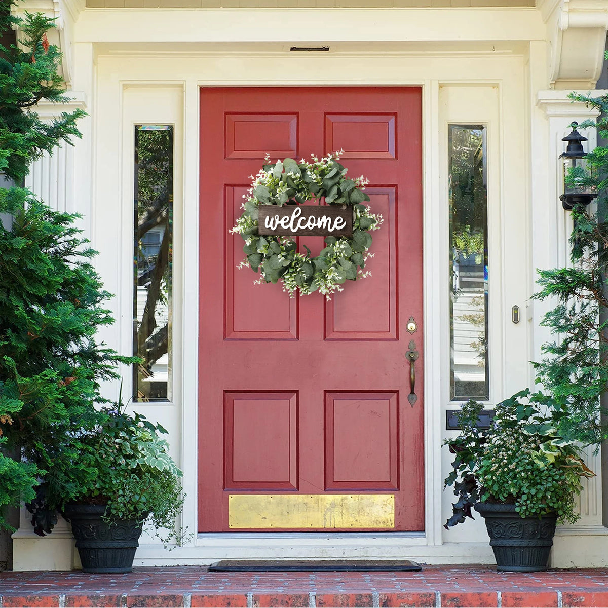 A red door decorated with an artificial eucalyptus wreath and a welcome sign, framed by white trim and greenery.