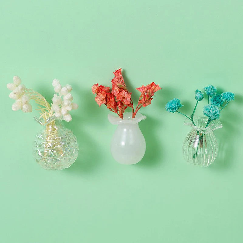 Three glass vases hold dried flowers in white, orange-red, and blue hues against a light green background.