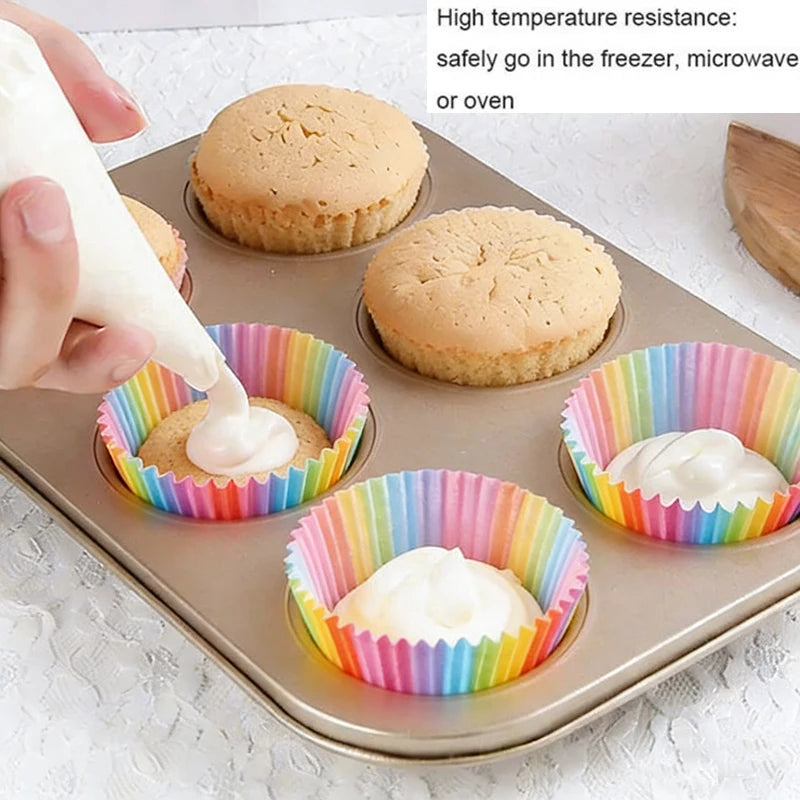 A tray of baked cupcakes in rainbow-colored disposable liners, with frosting being applied to some.