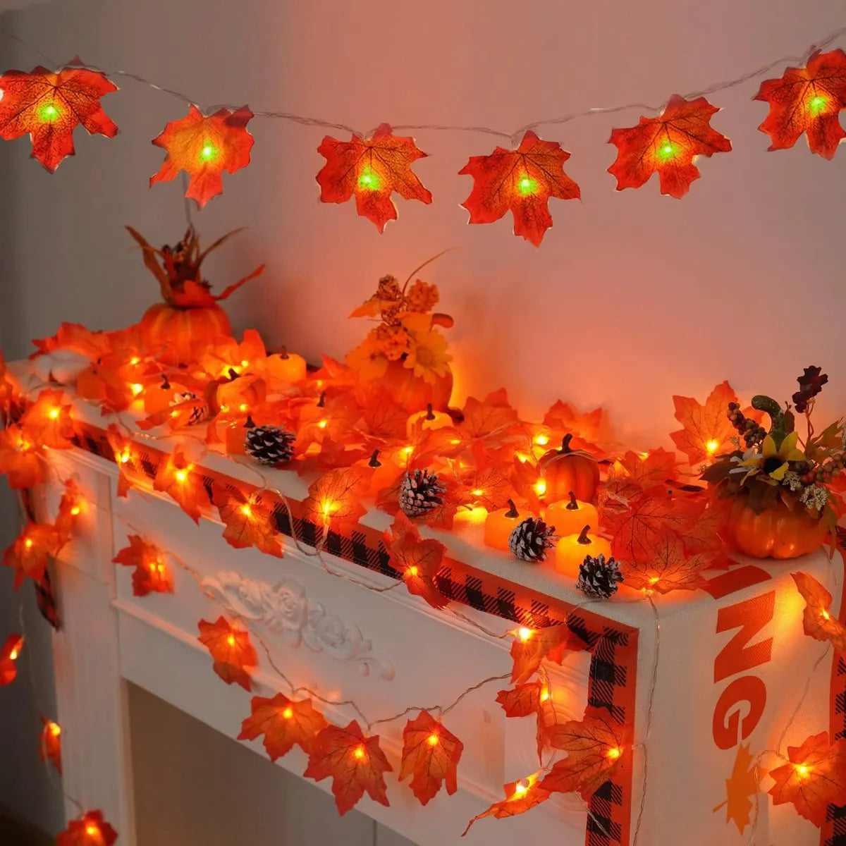 String lights shaped like autumn maple leaves illuminate a white fireplace mantel with orange and yellow warm light.