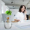 A woman works at a desk with an avocado seed growing kit in a glass vase on the table.