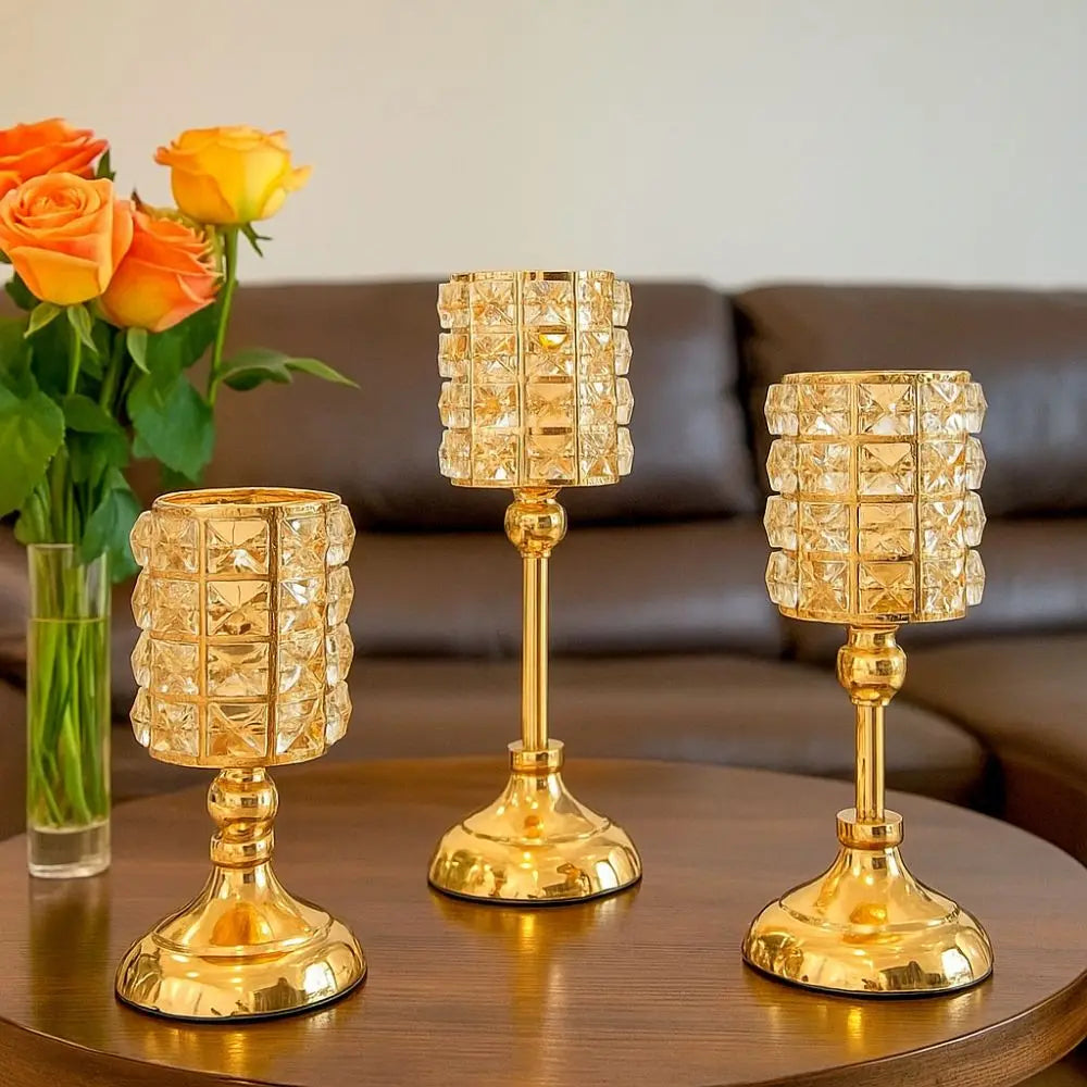 Un juego de tres candelabros de cristal dorado sobre una mesa de madera oscura con rosas naranjas de fondo.