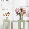 A light green ceramic vase holding pink flowers on a white table.