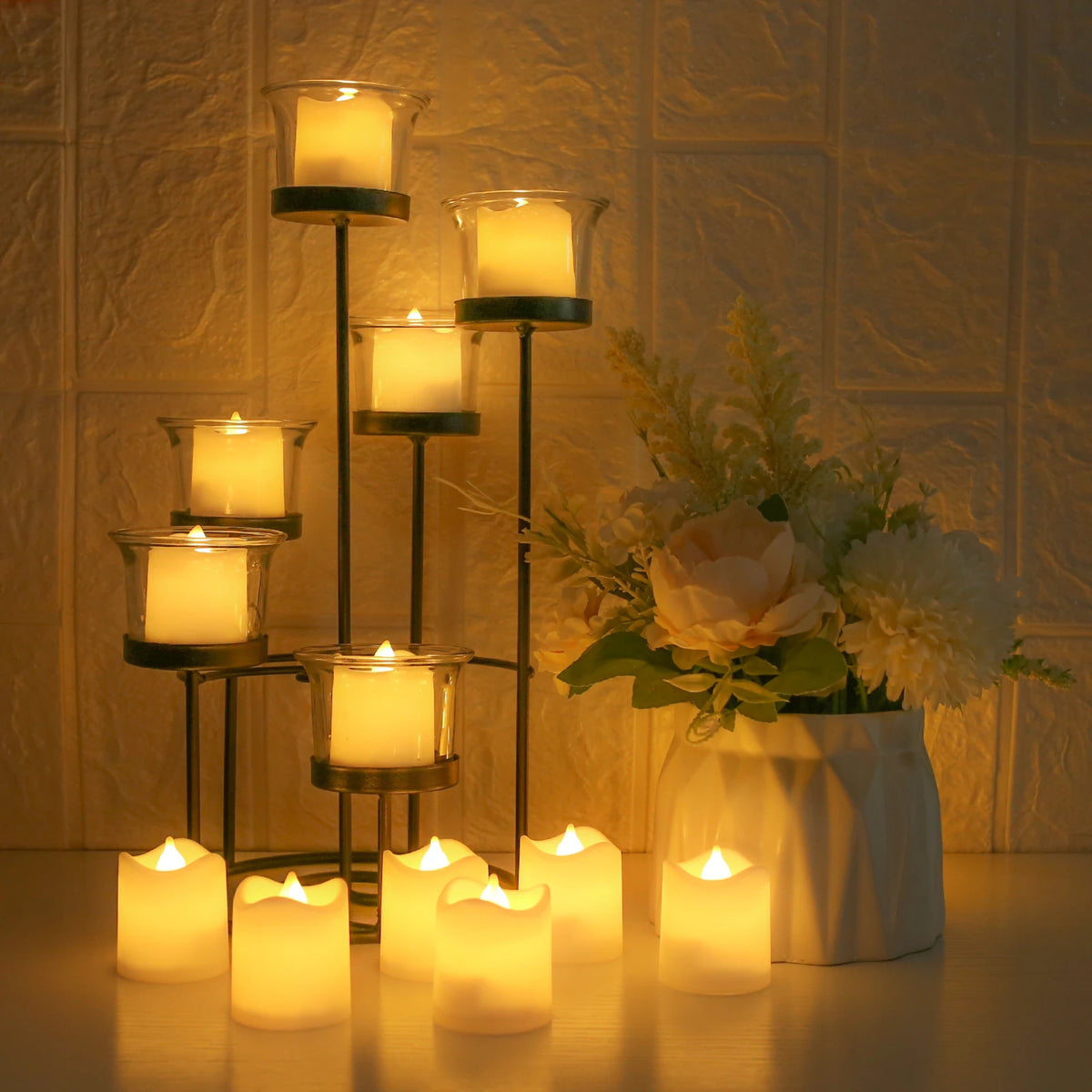 A collection of glowing LED votive tea lights displayed on a wooden surface with a floral arrangement in the background.