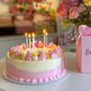 A pink and white layered cake decorated with floral frosting and lit birthday candles on a white platter.