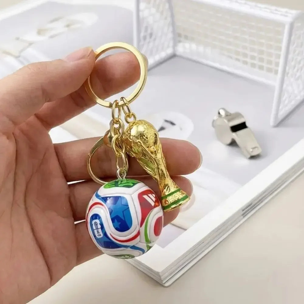 A hand holds a gold-colored FIFA trophy keychain featuring a soccer ball charm against a white background.
