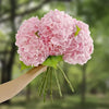 A hand holds a cluster of large pink artificial hydrangea flowers with green stems against a blurred green background.