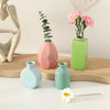 A collection of ceramic mini vases in pink, green, and blue colors with a pink floral arrangement on a white table.