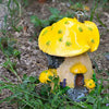 A resin mushroom house garden statue with a bright yellow cap and a small, textured body, set in natural soil and greenery.