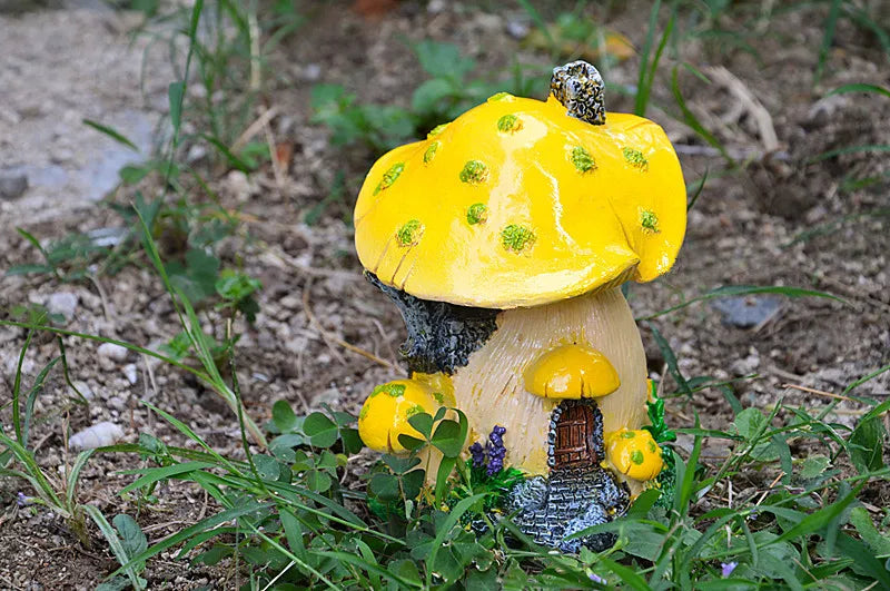 A resin mushroom house garden statue with a bright yellow cap and a small, textured body, set in natural soil and greenery.