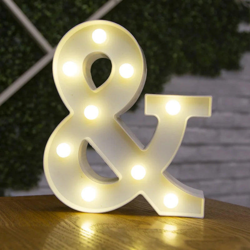A glowing warm white LED alphabet letter light shaped like an ampersand on a wooden surface.