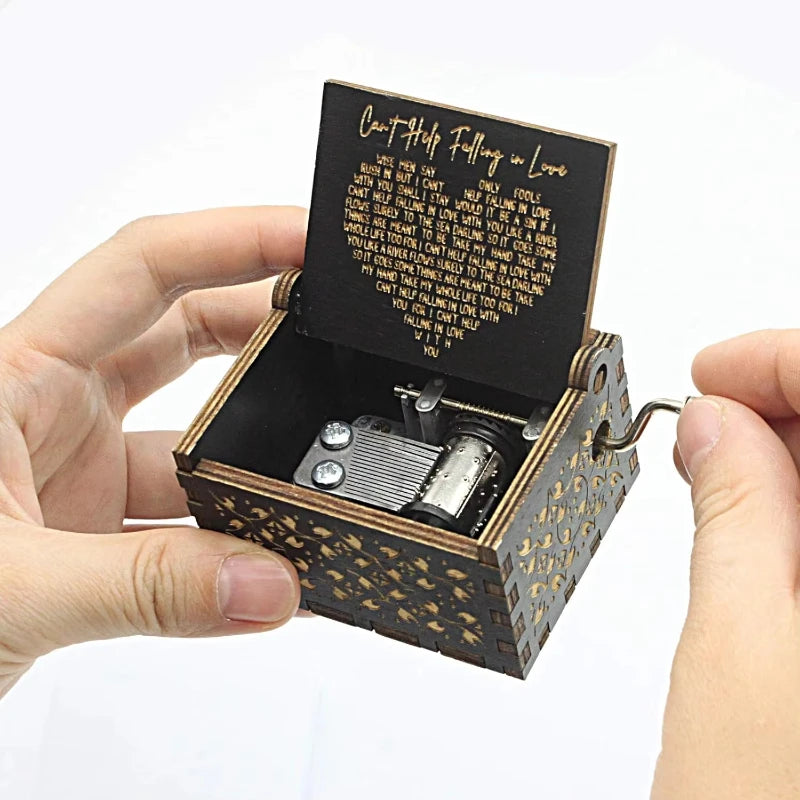An antique engraved wooden music box with a lid showing text and a winding key is held in a hand.