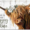 A close-up portrait of a Highland cow with horns and flowers, set against a rustic wooden background.