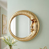 A round, golden crescent decorative wall mirror with a stylized face design mounted on a light green wall.