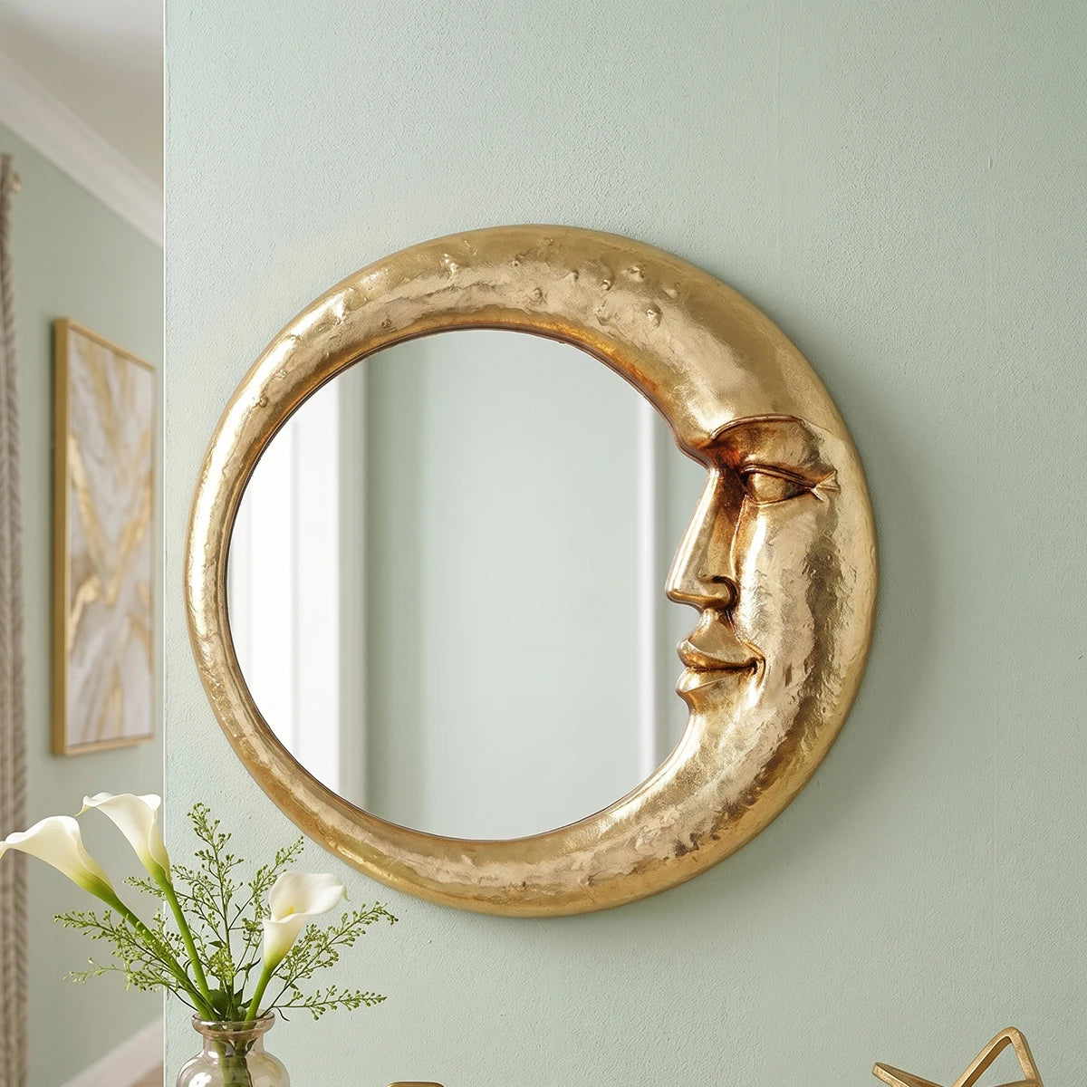 A round, golden crescent decorative wall mirror with a stylized face design mounted on a light green wall.