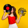 A smiling woman in a red top and white pants holds a black vinyl record and a circular wall clock with a heart design against