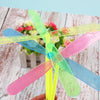 A hand holds a set of colorful bamboo dragonfly flying propeller toys with pink, blue, and green blades.