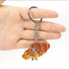 A silver acrylic keychain shaped like a sleeping cat on a croissant is held by a hand.