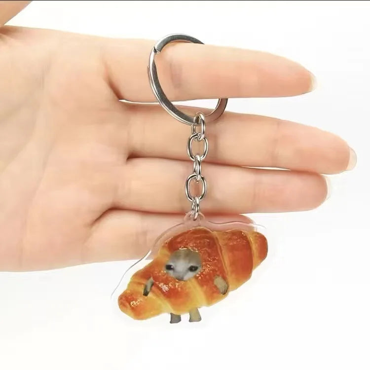 A silver acrylic keychain shaped like a sleeping cat on a croissant is held by a hand.