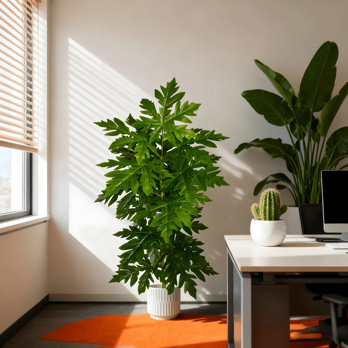 A vibrant green artificial papaya leaf plant in a white pot sits on a modern desk next to a computer.