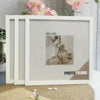 A set of three white wooden photo frames displayed on a light-colored surface with pink flowers in the background.