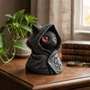 A dark gray gothic hooded cat statue with glowing red eyes sits on a wooden surface next to a stack of books.