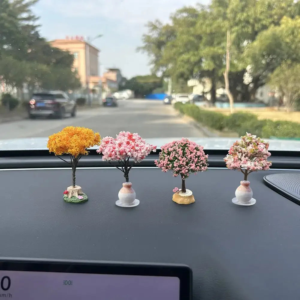 Four miniature tree ornaments with simulated pink and yellow flowers displayed on a car dashboard.