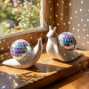 A white snail sculpture with a disco ball shell rests on a wooden surface near a sun catcher window.