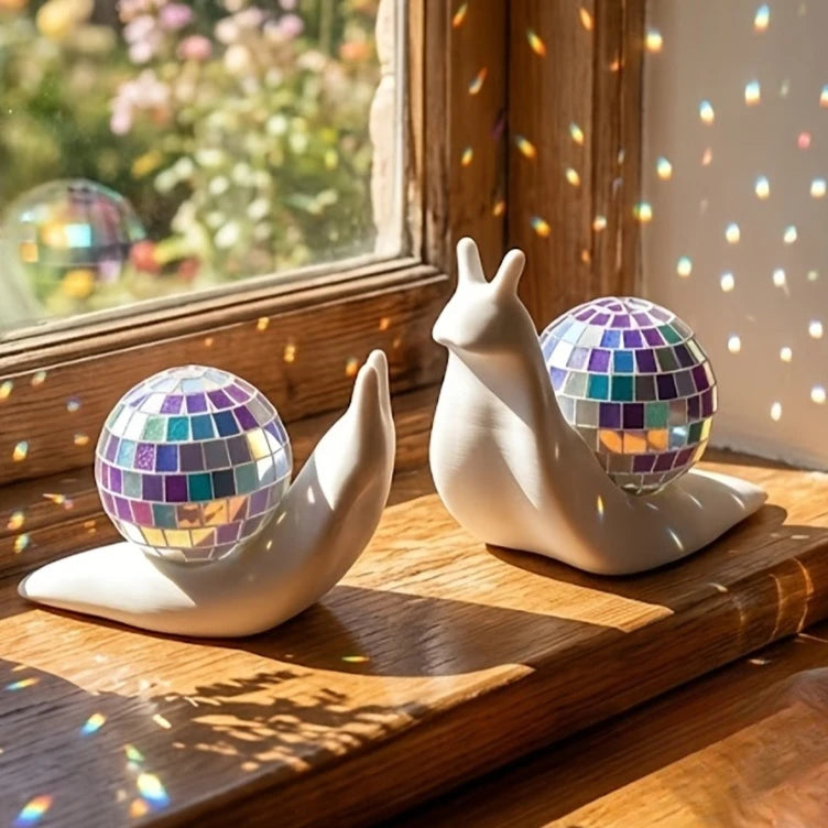 A white snail sculpture with a disco ball shell rests on a wooden surface near a sun catcher window.